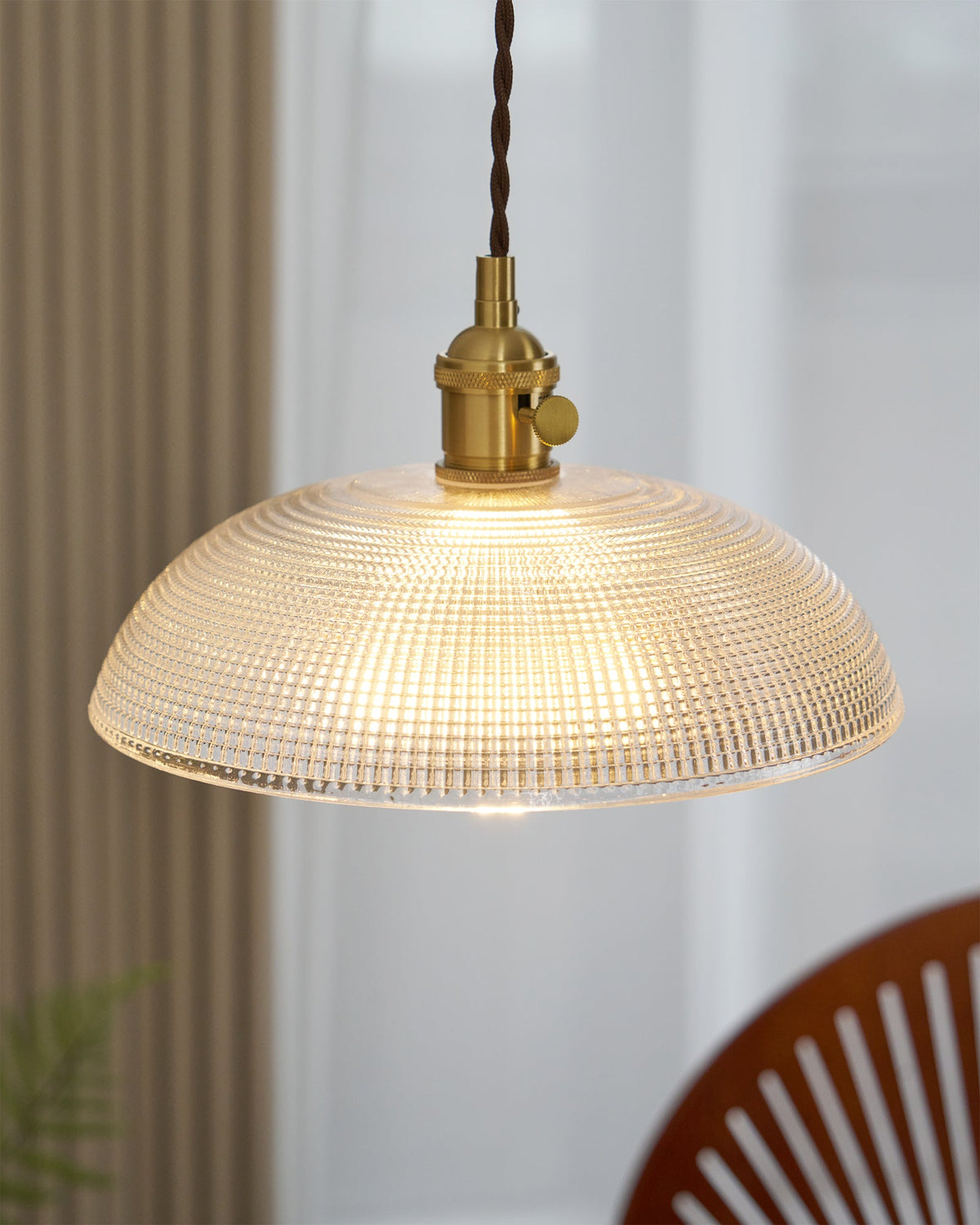 Near a curtained window, this Clérachic pendant hangs lit above a wood chair and a leafy plant.
