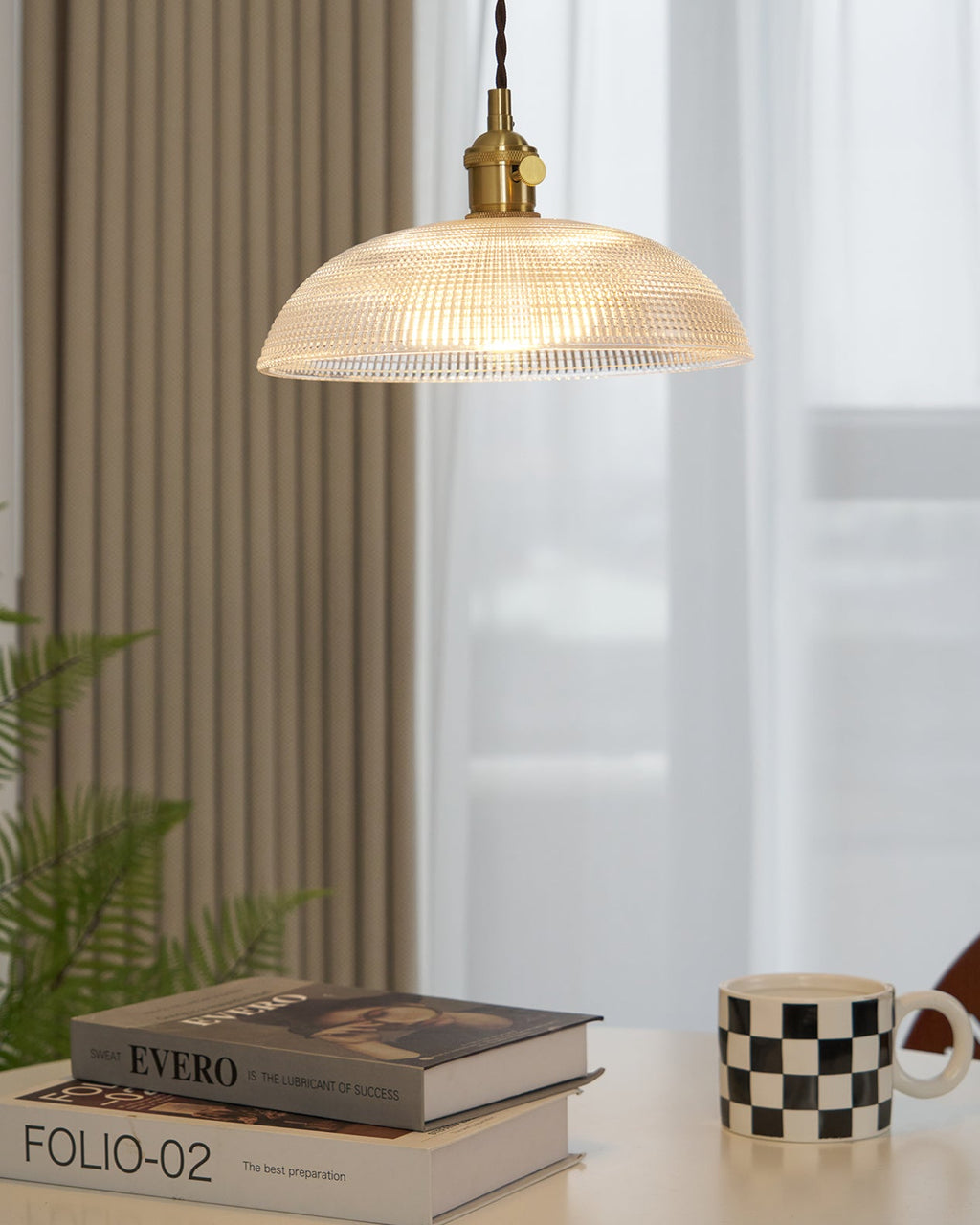 This Clérachic pendant glows above a table set with books and a chequered mug, near a curtained window.