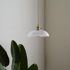 Unlit against a plain grey wall, this Clérachic pendant's ribbed glass shade sits beside a leafy plant.
