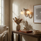 Above a console table beside an upholstered armchair, this Clérachic sconce's nickel S-curve holds two glowing alabaster globes.