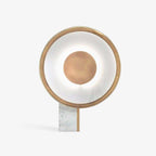 Clérachic gold-ringed disc table lamp on a plain white background, its rectangular white marble foot standing below the softly lit ring.