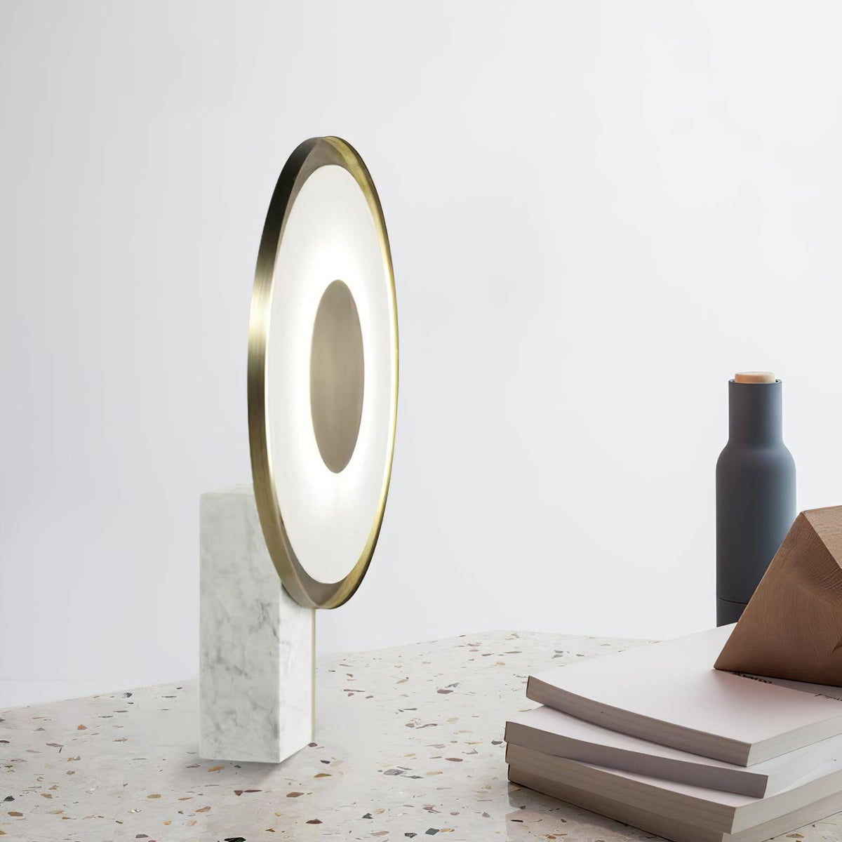 Clérachic lamp glowing on a terrazzo floor beside a dark bottle-shaped vase and a stack of books nearby.