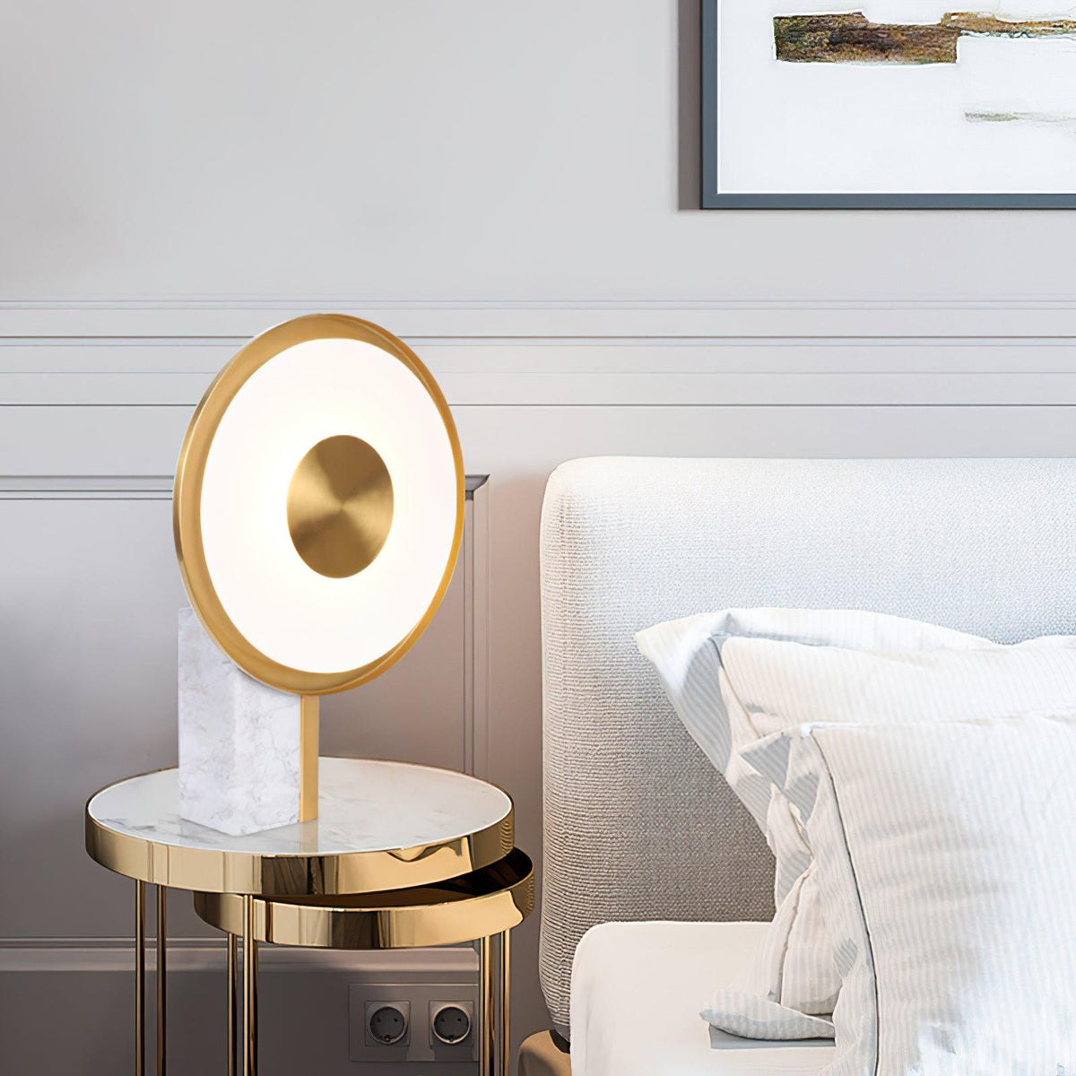 A Clérachic table lamp stands on a round marble-top side table next to a cream armchair with a bolster cushion, gold legs beneath.