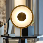 The Clérachic disc lamp glows on a round black side table, its gold rim catching the light against a softly blurred blue room.
