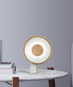 Clérachic disc lamp glowing on a small dark side table against a plain grey wall, a stack of books resting beside its marble foot.