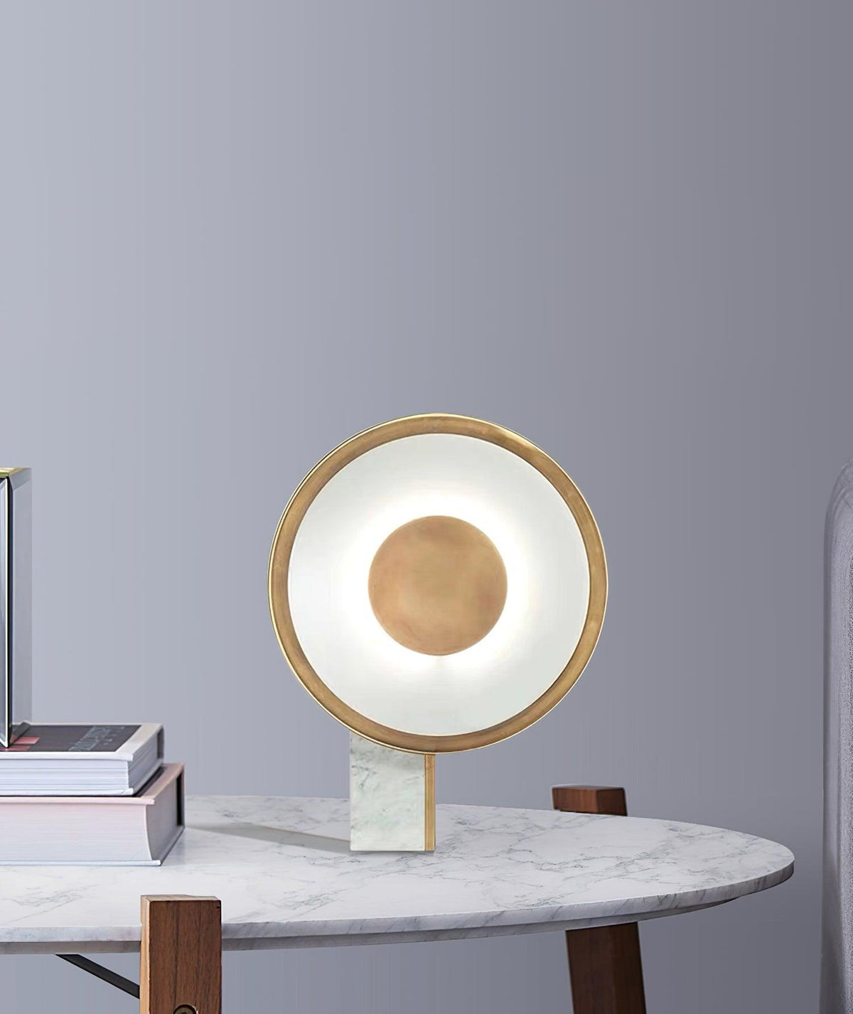Clérachic disc lamp glowing on a small dark side table against a plain grey wall, a stack of books resting beside its marble foot.