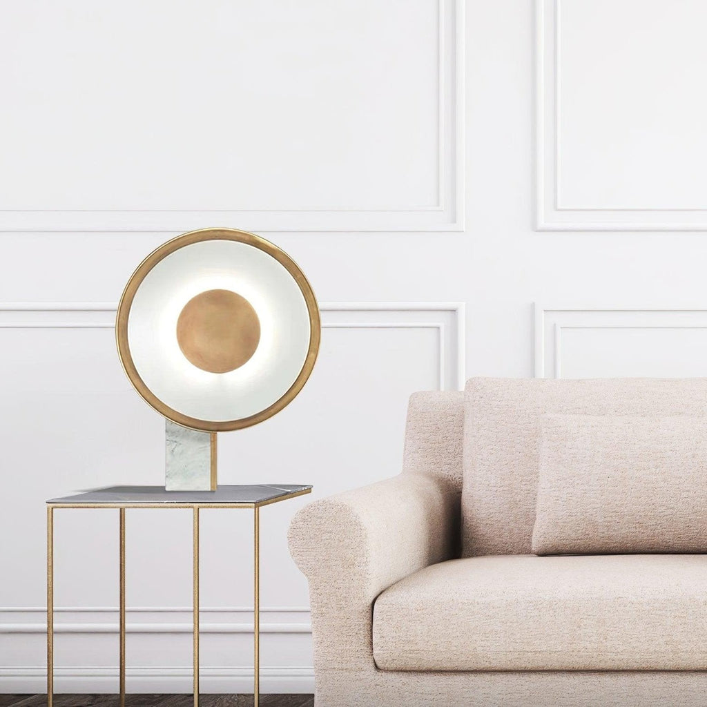Clérachic table lamp lit on a gold-framed marble side table beside a beige linen sofa, set against a panelled white wall.
