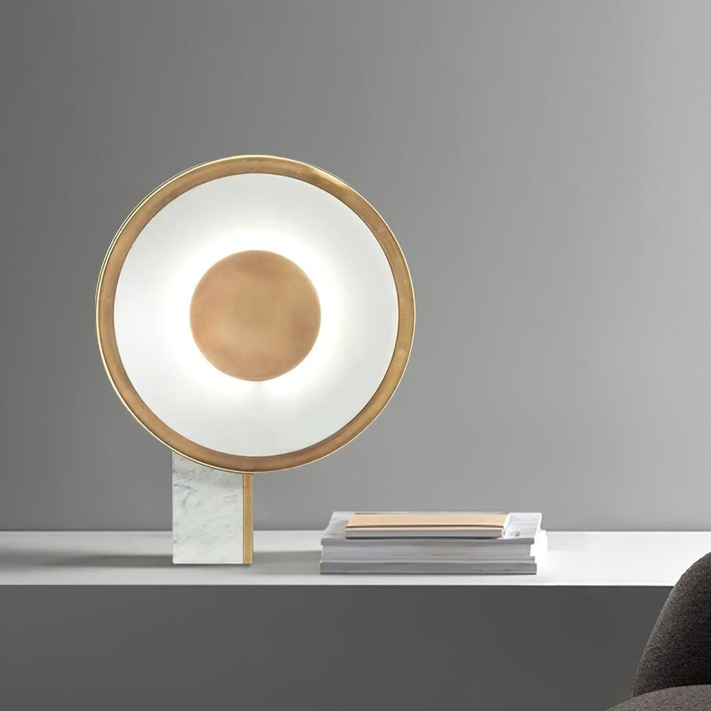 Clérachic gold disc lamp lit on a white marble-top table against a grey background, a stack of magazines resting beside it.