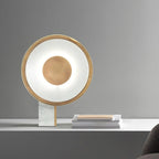 Clérachic gold disc lamp lit on a white marble-top table against a grey background, a stack of magazines resting beside it.