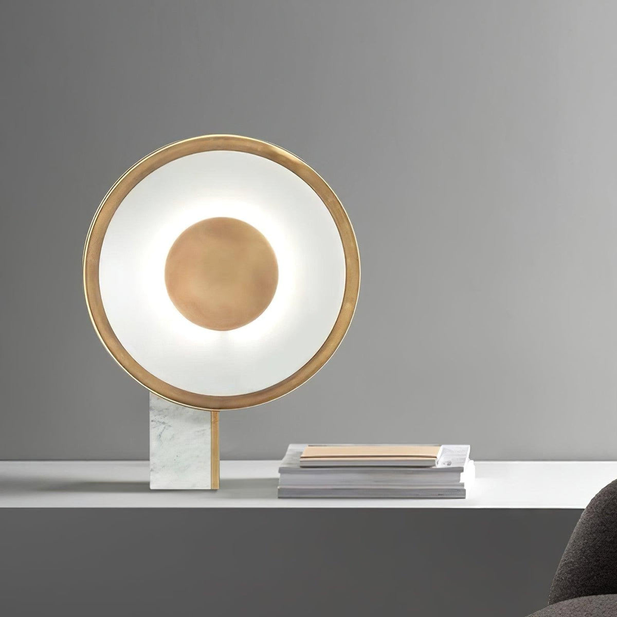 Clérachic gold disc lamp lit on a white marble-top table against a grey background, a stack of magazines resting beside it.