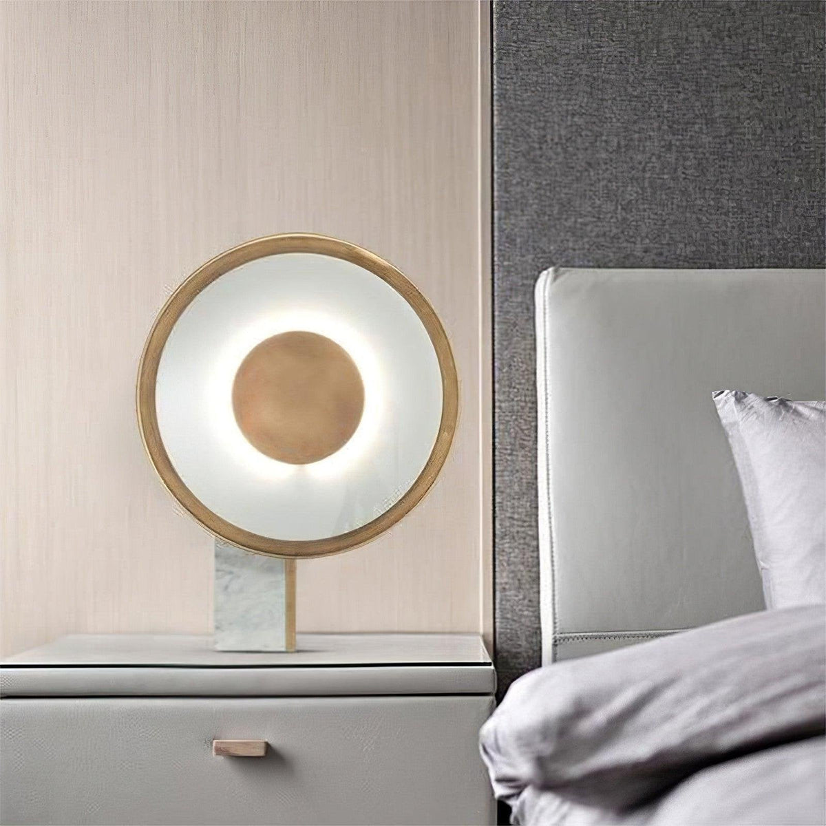 The Clérachic disc lamp stands on a bedside table against a light wood panel wall, beside a grey upholstered headboard.