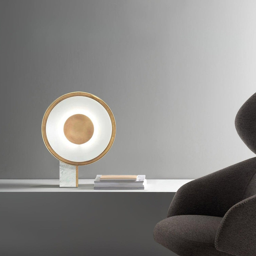 Clérachic table lamp lit on a marble table against a grey background, the edge of a dark armchair and stacked magazines nearby.