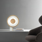 Clérachic table lamp lit on a marble table against a grey background, the edge of a dark armchair and stacked magazines nearby.