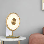Clérachic gold disc table lamp on a marble side table, a dusty pink armchair edge and plain grey wall filling the background.