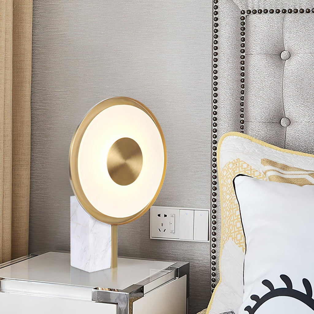Clérachic gold disc lamp lit on a bedside table beside a tufted grey headboard, a wall switch plate visible just behind it.