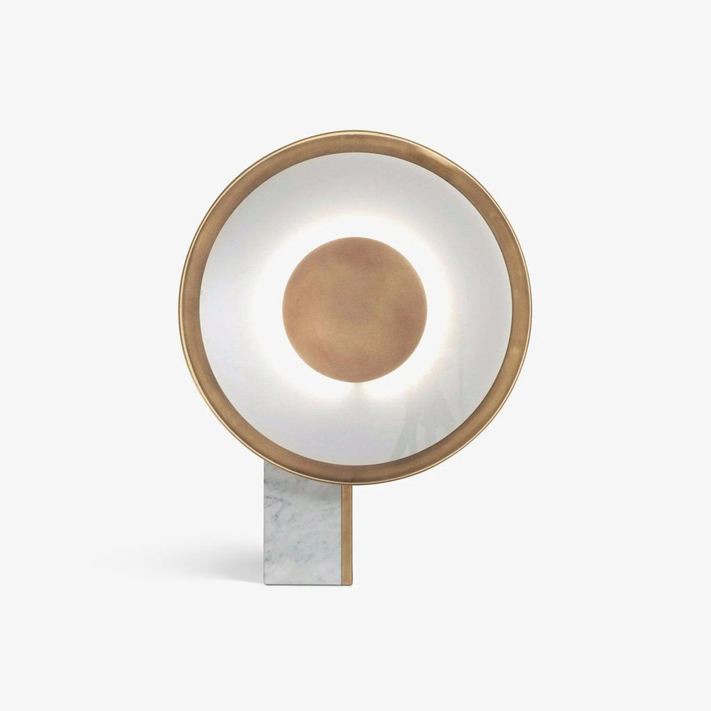 Clérachic gold disc table lamp shown straight on, lit, its rectangular marble foot planted on a plain background.
