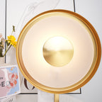 Extreme close-up of the Clérachic lamp's gold rim and brushed centre disc, with flowers just visible at the frame's edge.