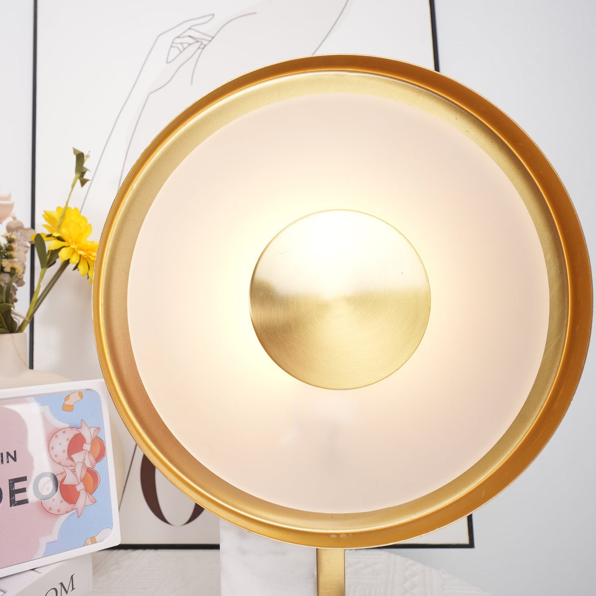 Extreme close-up of the Clérachic lamp's gold rim and brushed centre disc, with flowers just visible at the frame's edge.