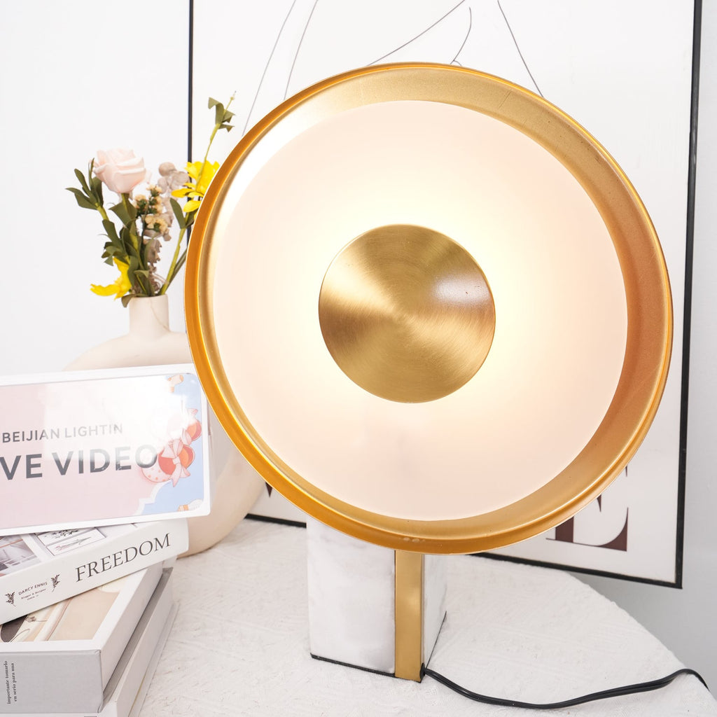 Clérachic disc lamp glowing warm beside a rose in a vase and stacked books, a line-art figure print in the background.