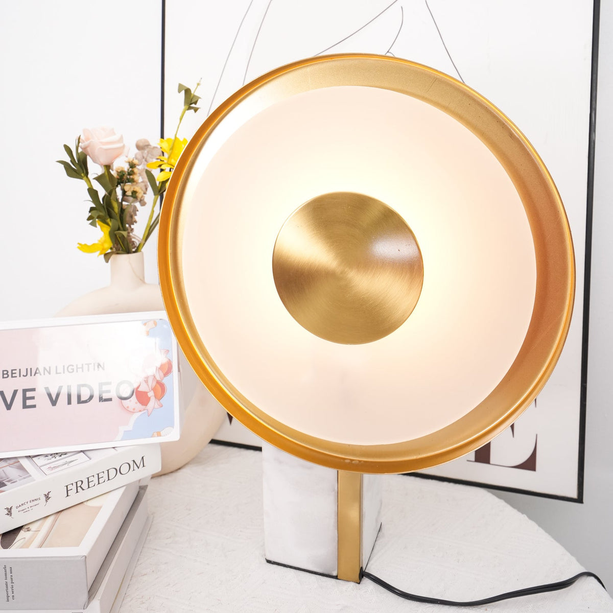Clérachic disc lamp glowing warm beside a rose in a vase and stacked books, a line-art figure print in the background.