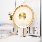 Clérachic disc lamp lit warm beside flowers and books, its brushed-gold centre bright against the softly glowing ring.