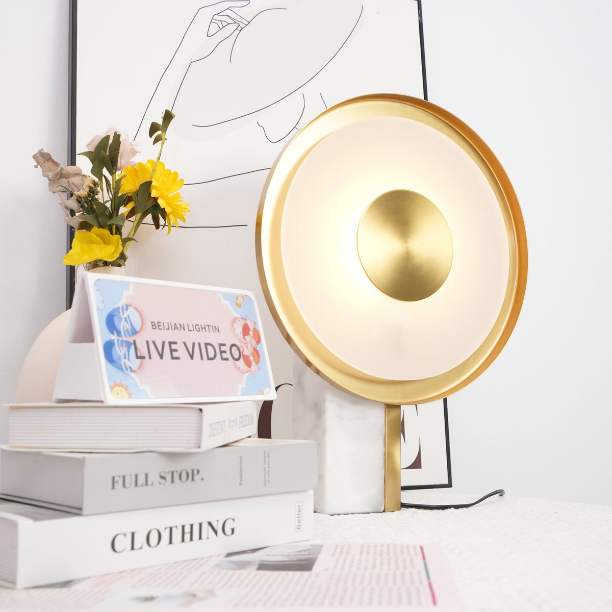 Clérachic disc lamp glowing beside yellow flowers and stacked books titled Full Stop and Clothing, a line-art print behind it.