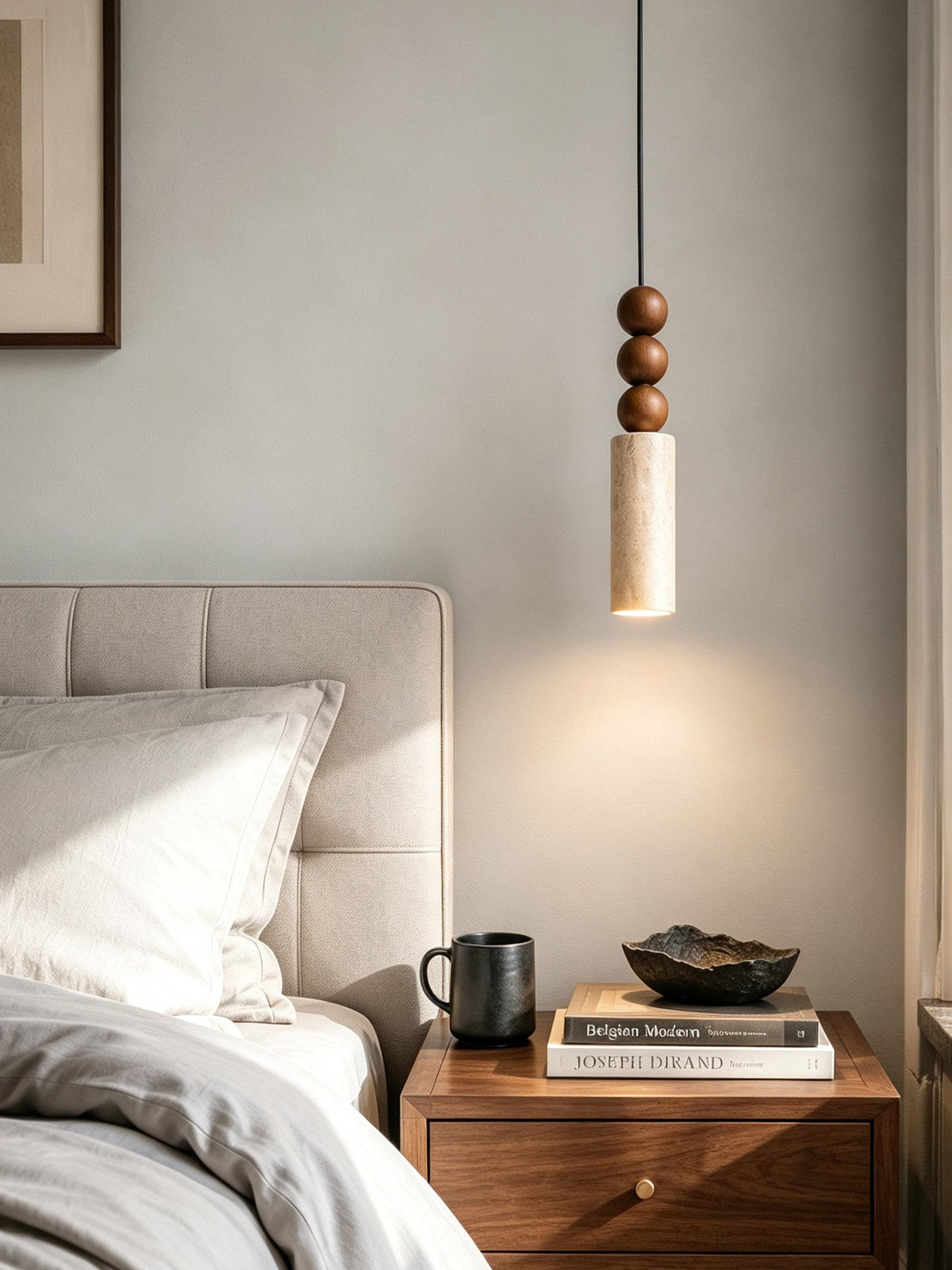 This Clérachic pendant glows above a nightstand set with a mug, stacked books and a small ceramic bowl, beside a padded headboard.