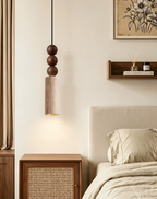 This Clérachic pendant hangs beside a bed with a woven cane nightstand, its travertine cylinder glowing warm beneath stacked wood beads.