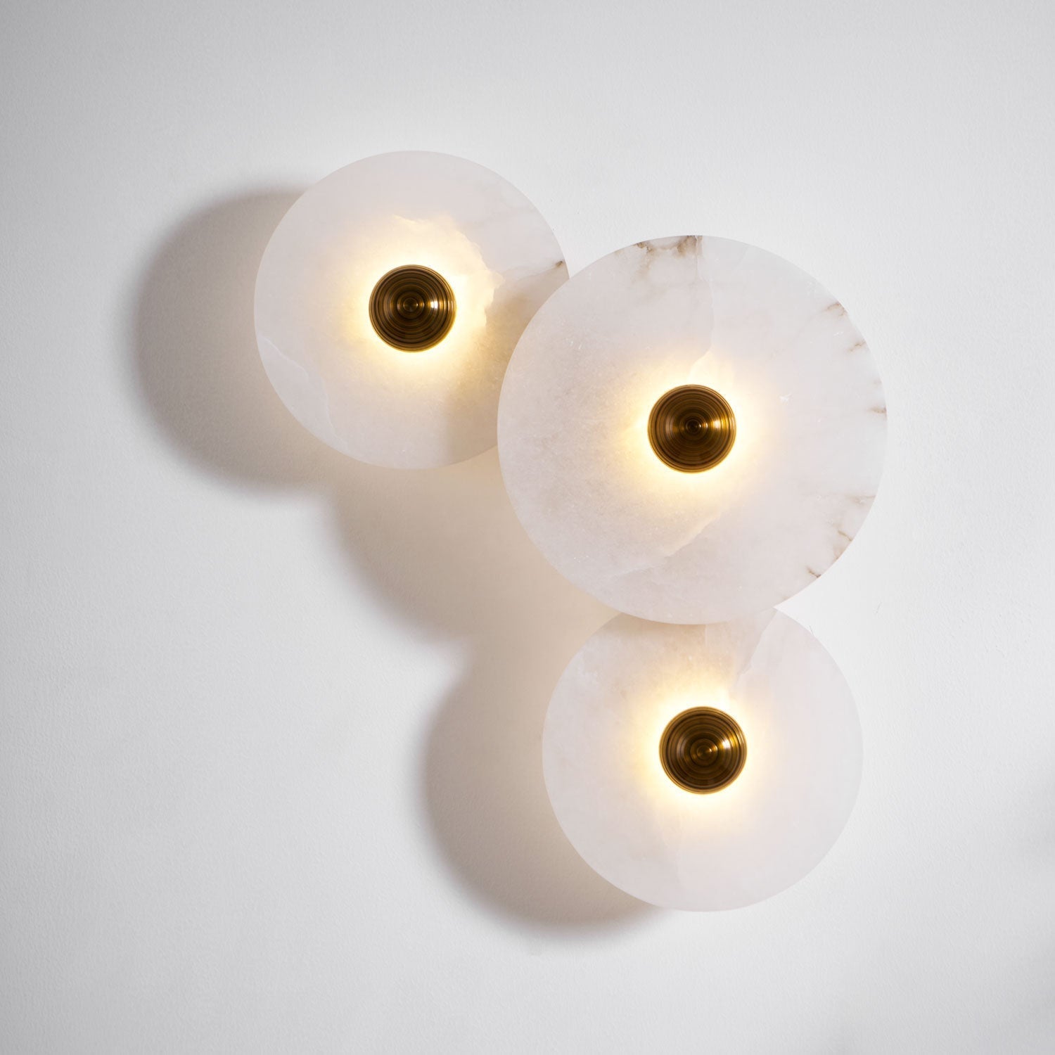 Three lit alabaster discs on a white wall, brass centres bright and shadows cast to the left.