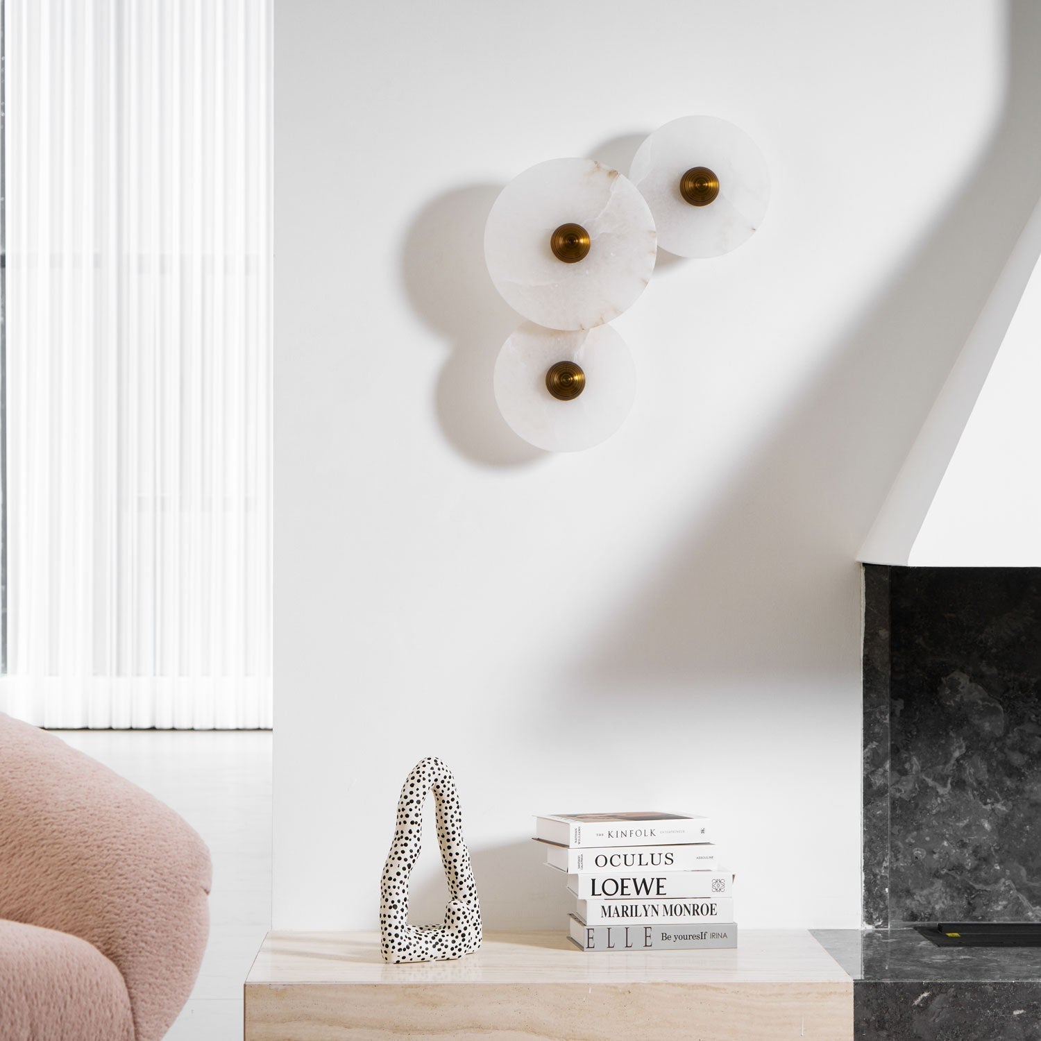 Three unlit alabaster discs on a white wall above a bench with books and a spotted sculpture.