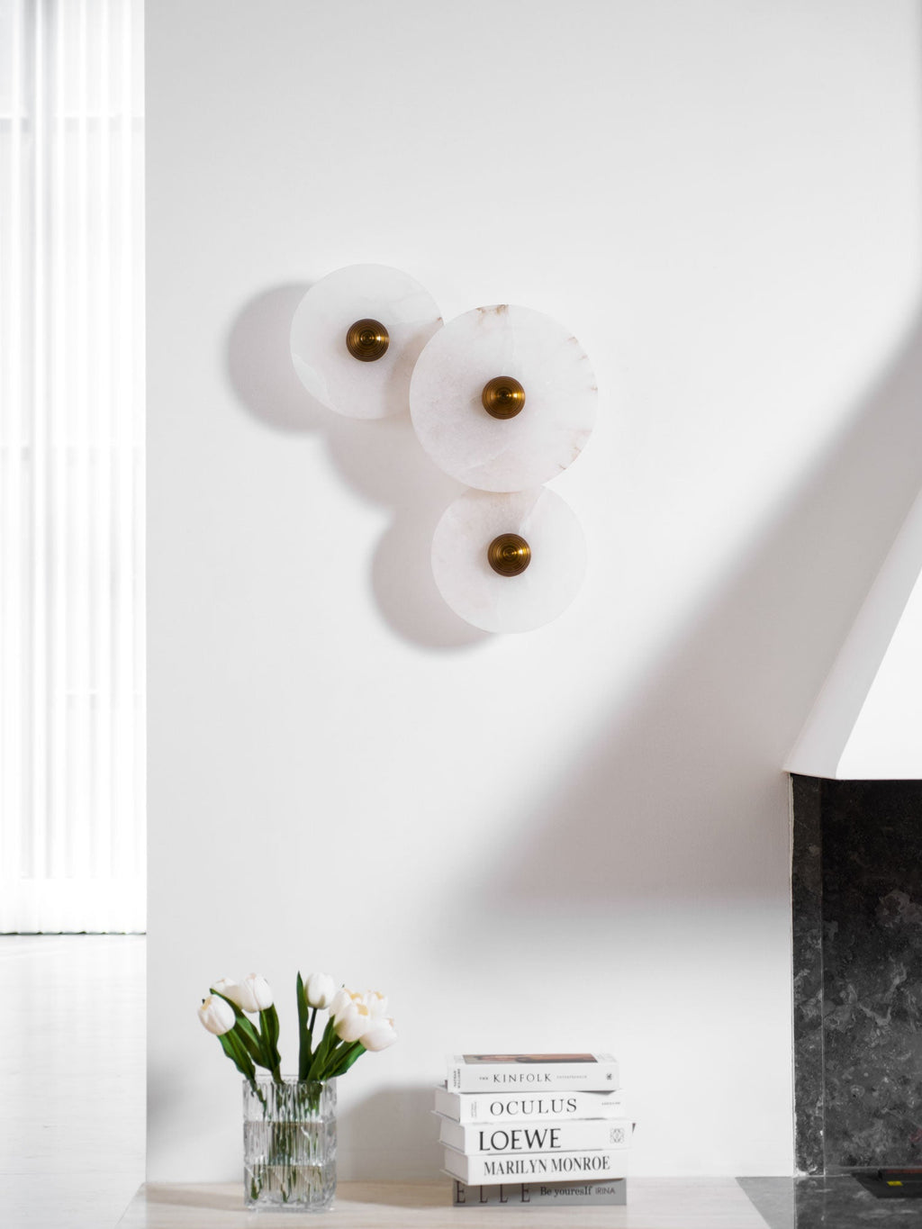 Unlit three-disc cluster on a sunlit white wall, with white tulips on the ledge below.