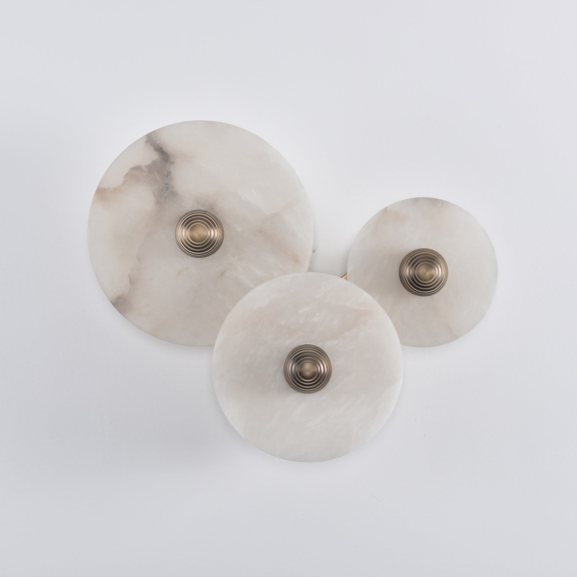 Three unlit alabaster discs on a white wall, brass bosses catching light, stone veining in shadow.