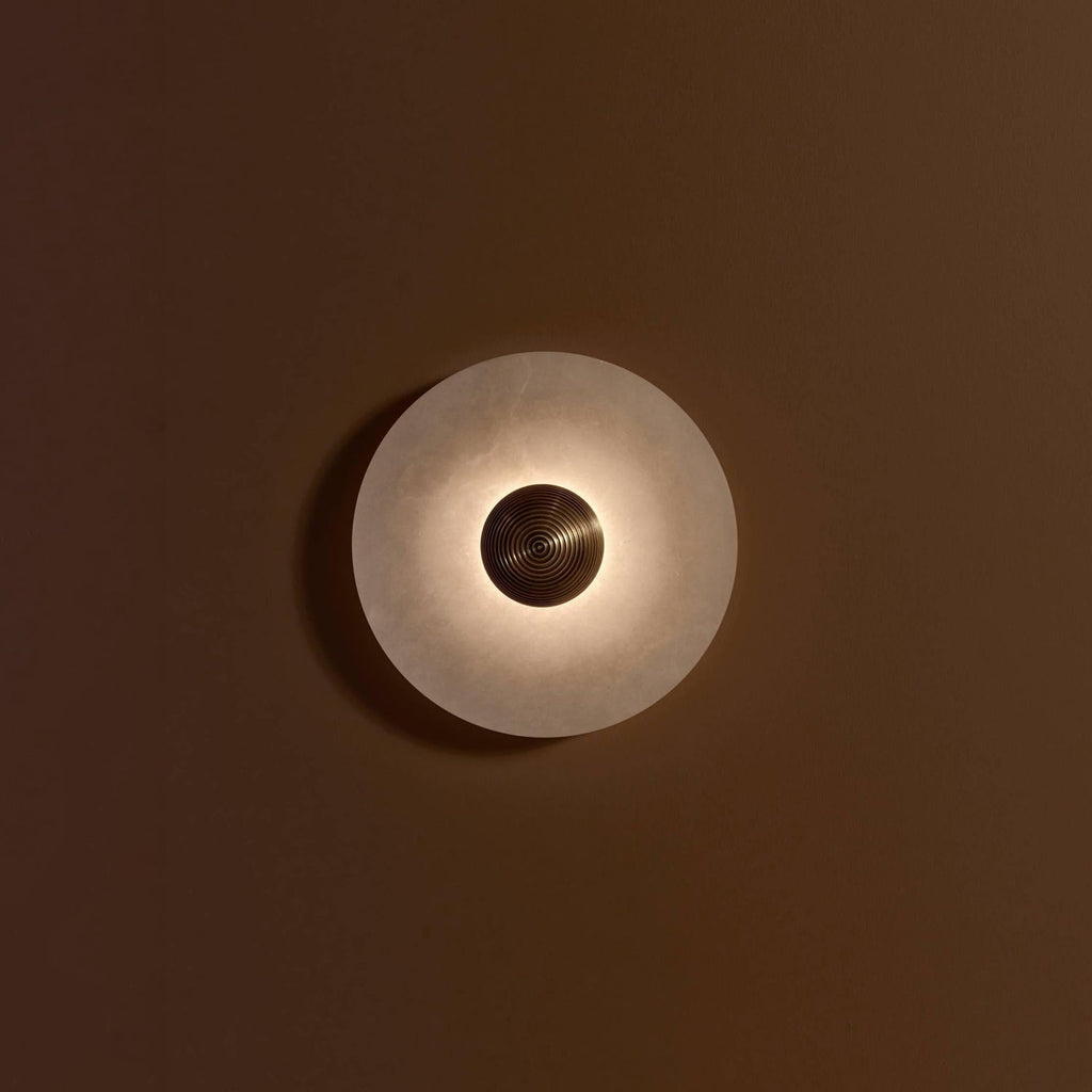 A Clérachic single sconce lit on a brown wall, light spilling in a ring around the ribbed gold centre.