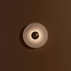 A Clérachic single sconce lit on a brown wall, light spilling in a ring around the ribbed gold centre.