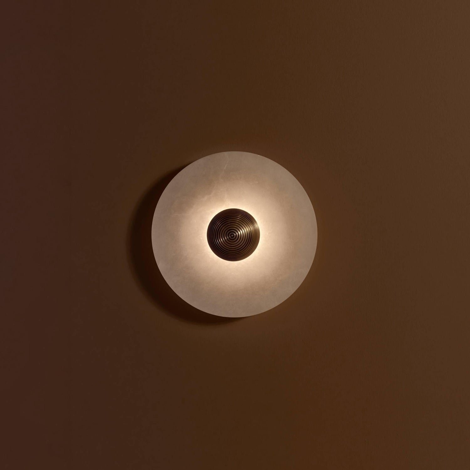 A Clérachic single sconce lit on a brown wall, light spilling in a ring around the ribbed gold centre.
