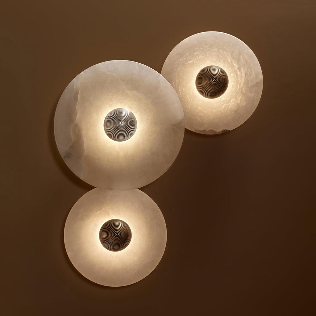 Three Clérachic lit sconces on a chocolate wall, the alabaster glowing translucent around bronze and steel bosses.
