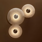 Three Clérachic lit sconces on a chocolate wall, the alabaster glowing translucent around bronze and steel bosses.