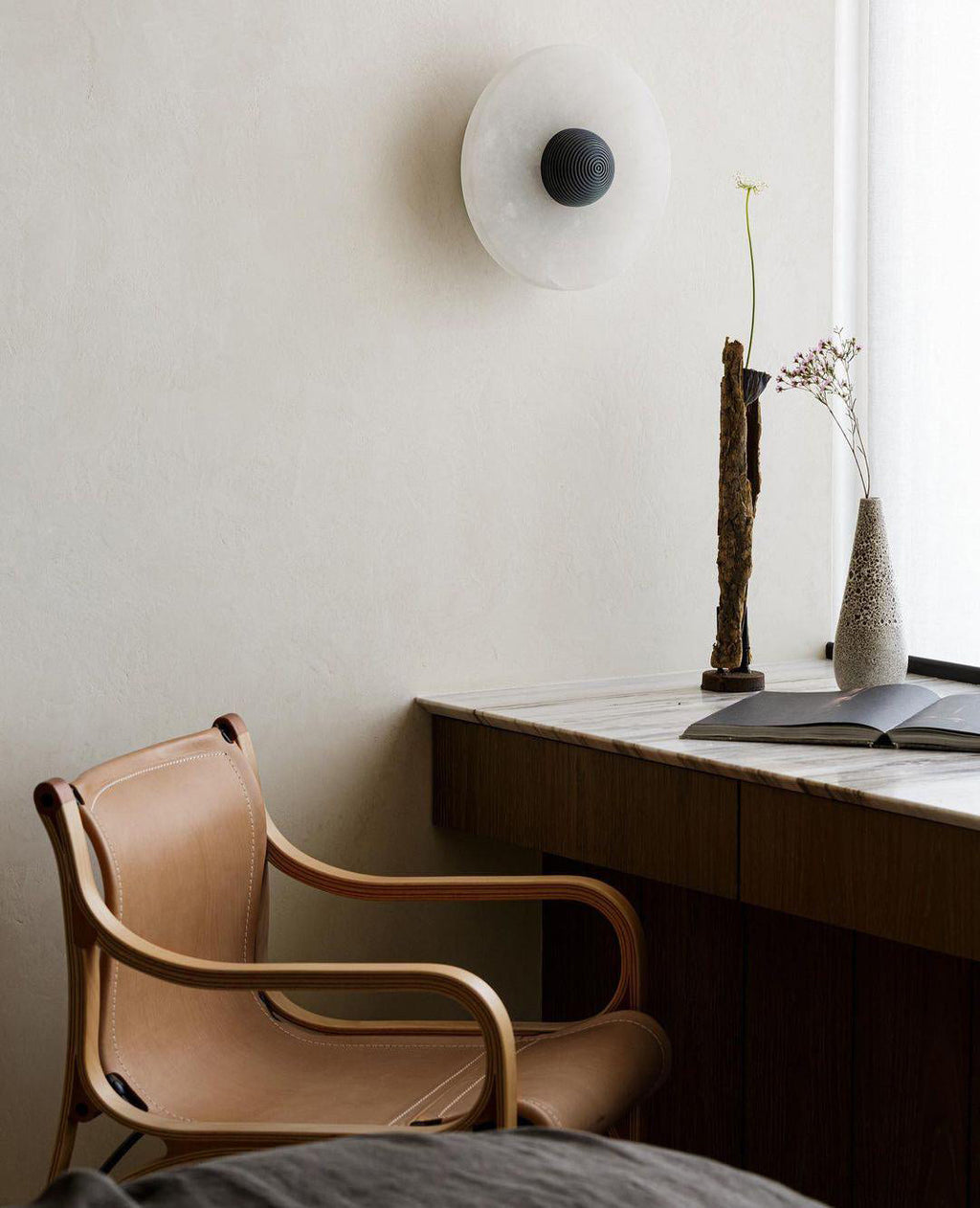 A Clérachic single sconce with a dark ribbed centre on a cream plaster wall above a marble desk and leather chair.