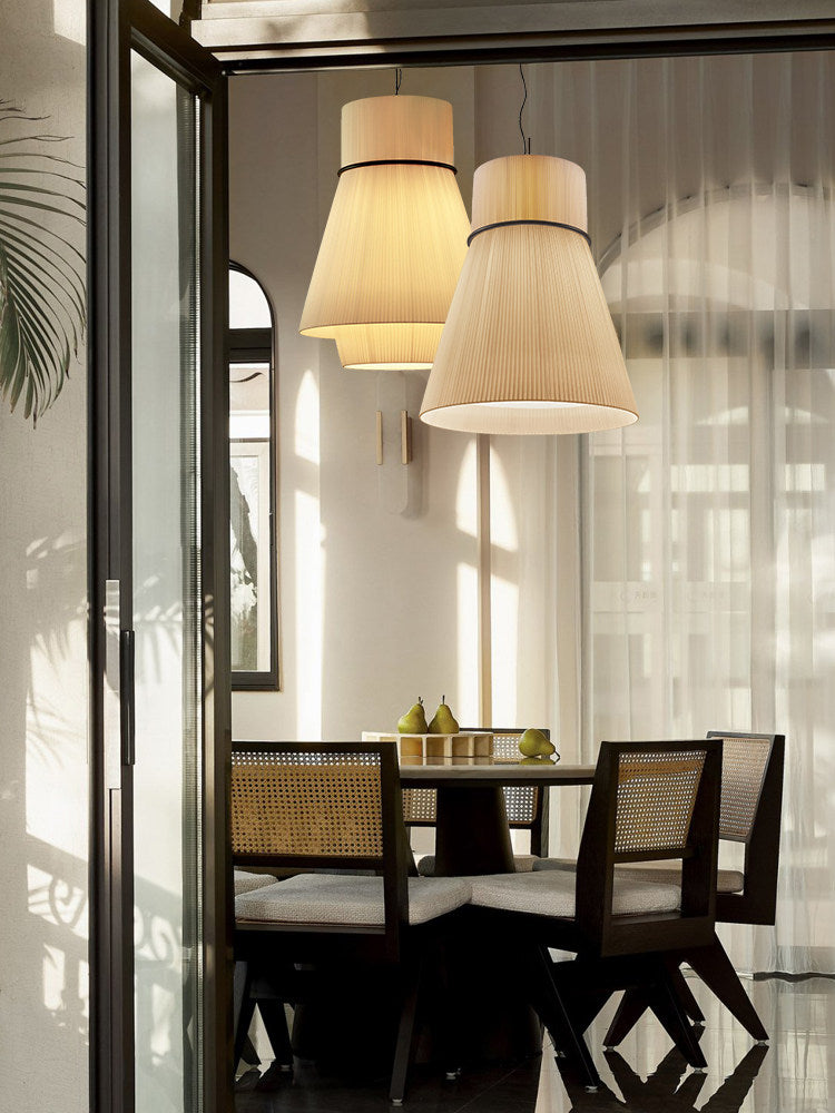 Two pleated Clérachic pendants glow side by side in a sunroom with glass doors and rattan chairs.