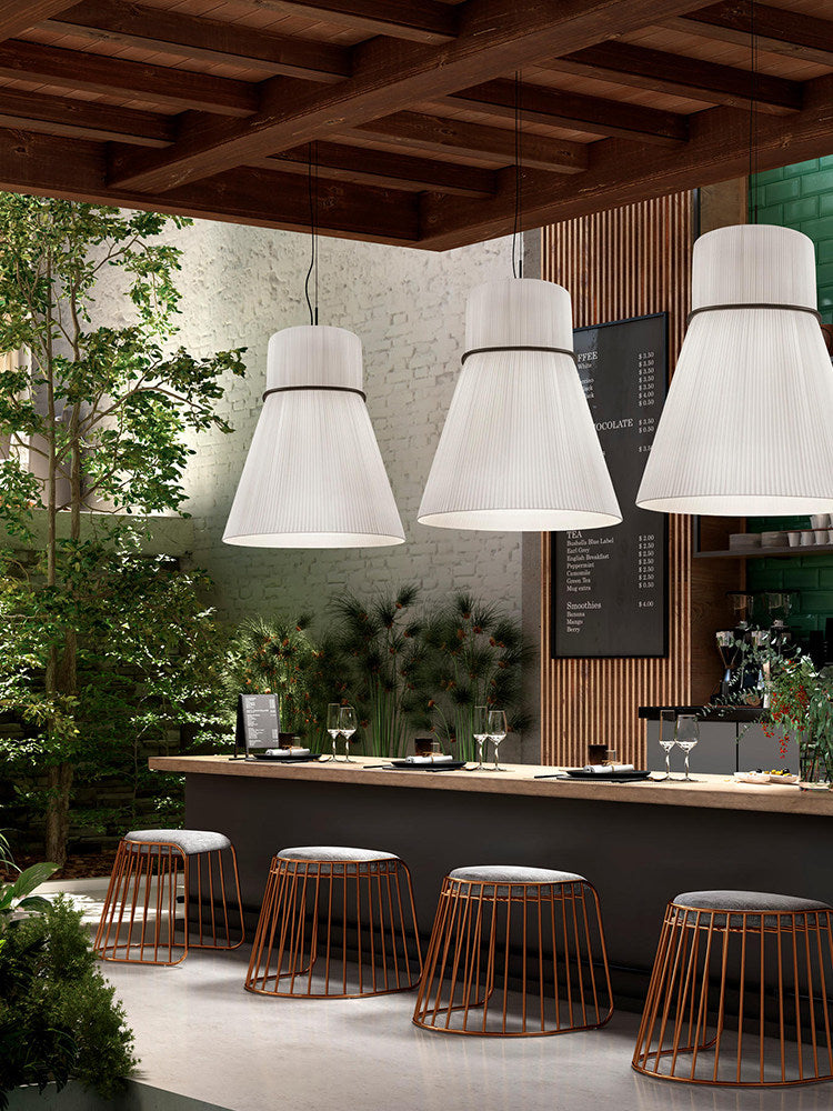 Three pleated Clérachic pendants light an outdoor bar counter lined with woven stools and potted greenery.