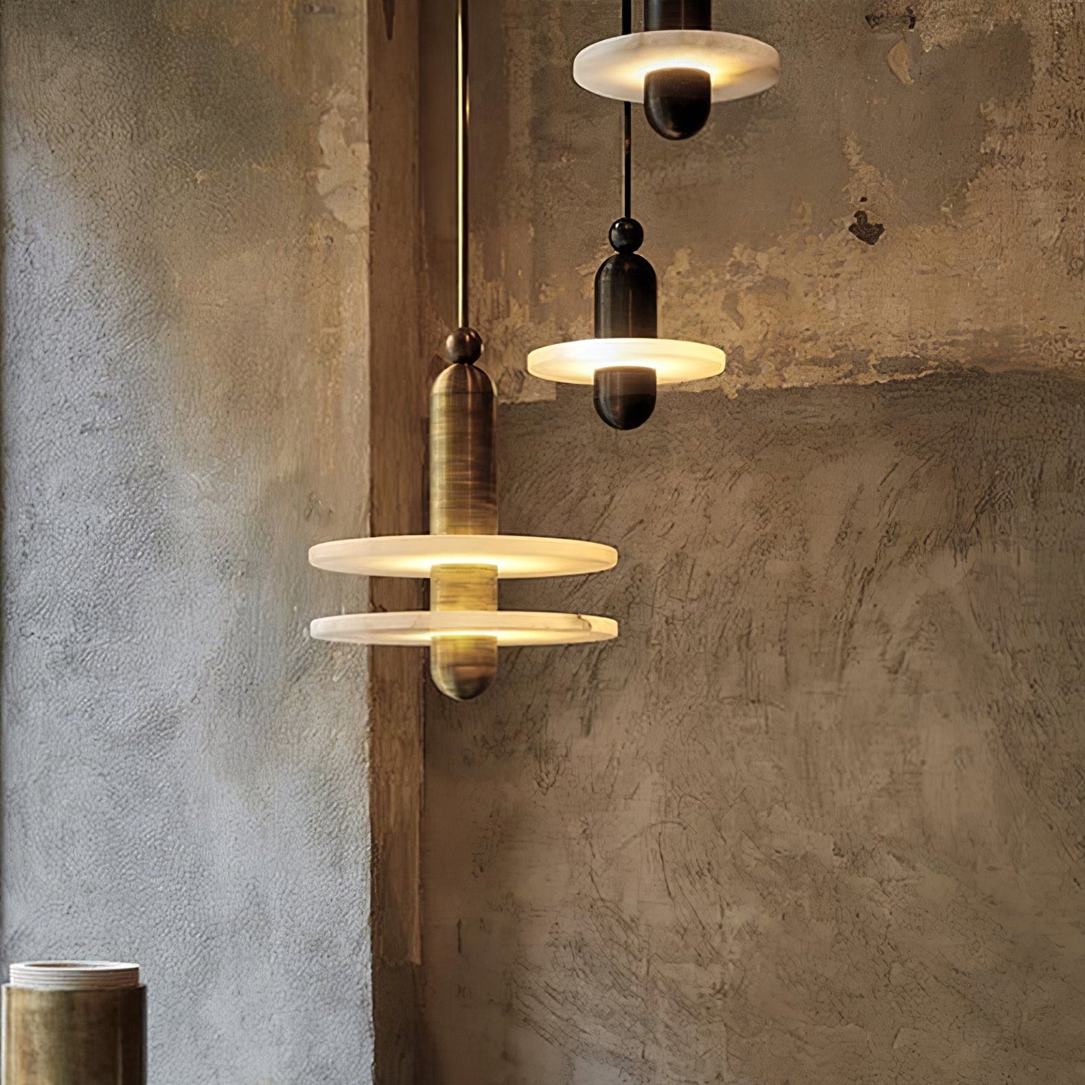 Three Orion pendants lit above a rustic plaster wall, mixing the gold two-disc and black one-disc forms.