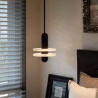Black two-disc Orion pendant lit beside a bed, next to a window with slatted blinds.