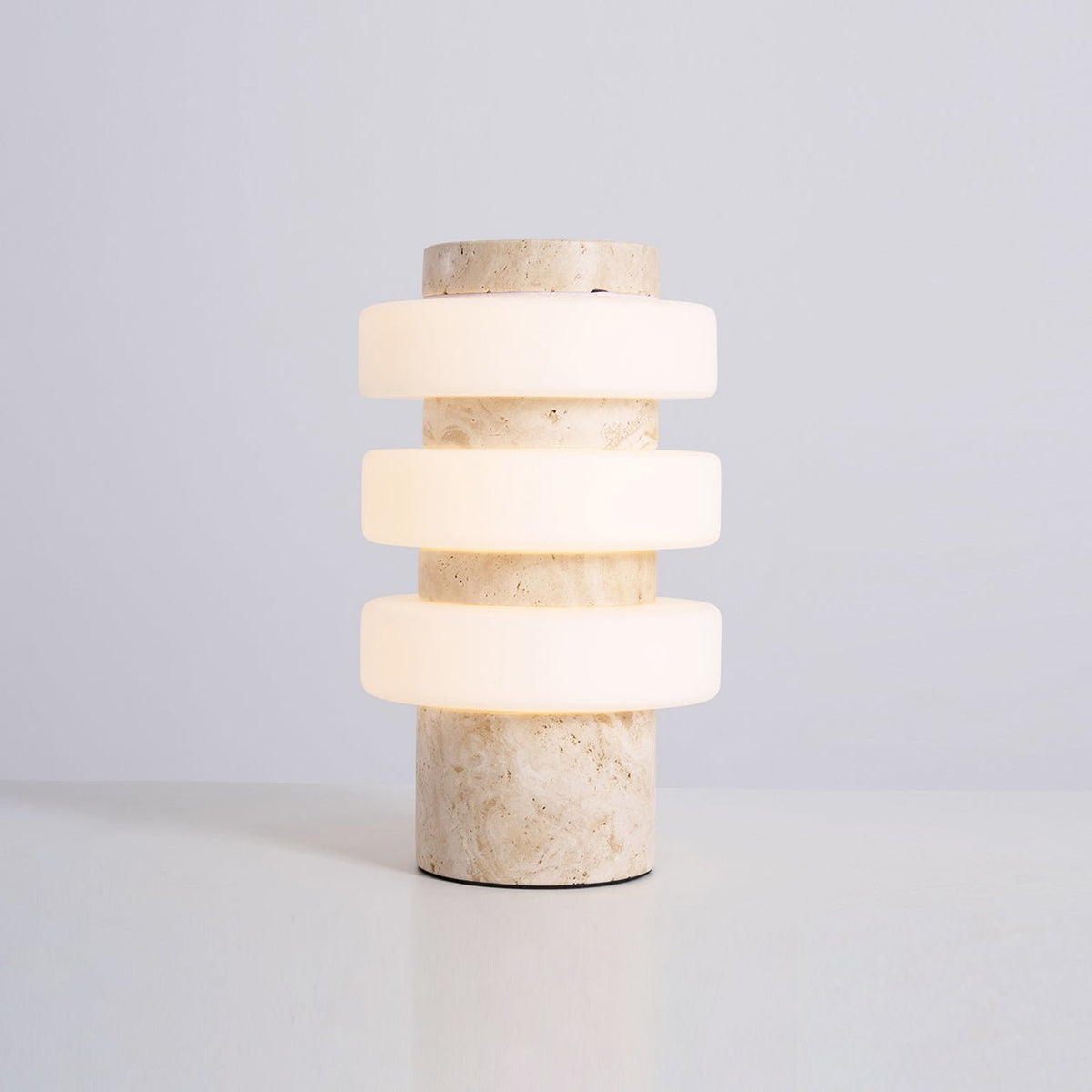 Studio shot of the lit Clérachic travertine table lamp on a plain grey background, its three glass rings glowing.