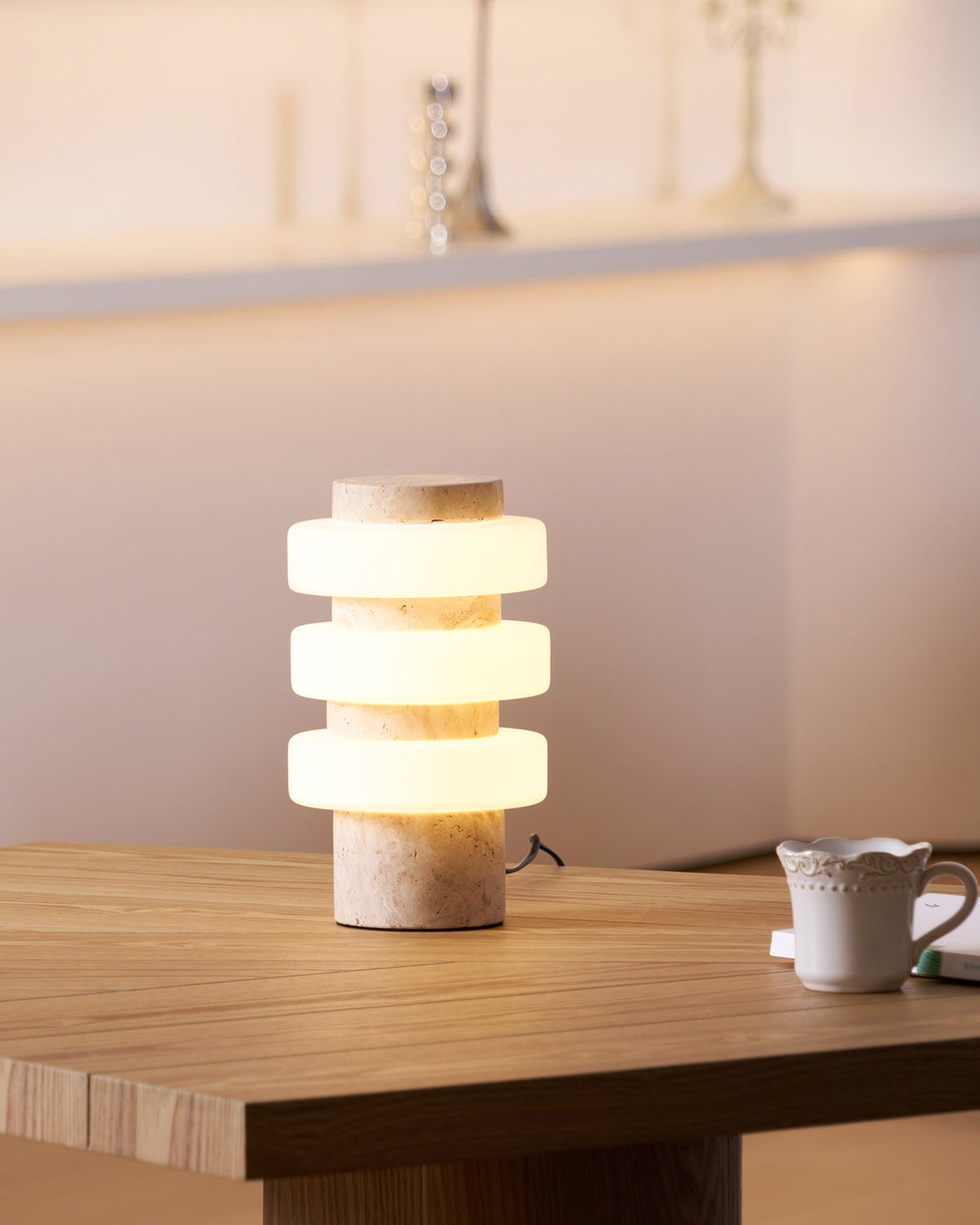 Clérachic travertine table lamp lit on a wood dining table, its three glass rings softly aglow beside a small jug.