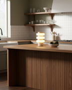 Clérachic travertine table lamp lit on a wood kitchen island, with open shelves and a tiled backsplash behind it.