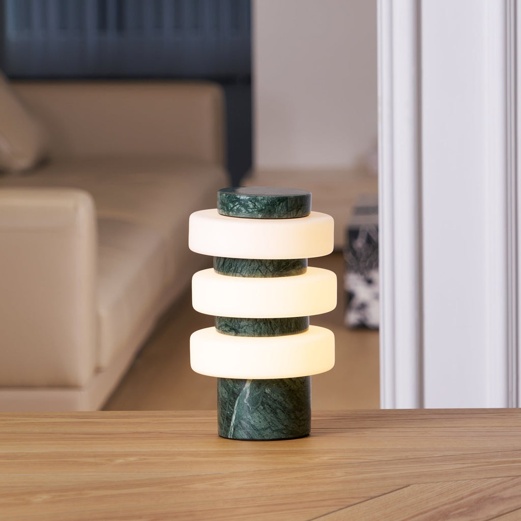 Clérachic green marble table lamp lit on a wood table in a living room, a pale sofa softly blurred behind it.