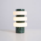 Studio shot of the lit Clérachic green marble table lamp on a white background, its glass rings glowing warmly.