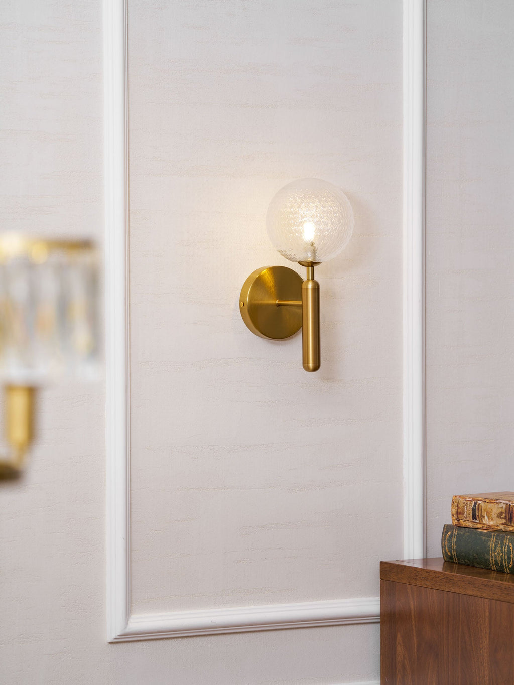Near a stack of books and a wood cabinet, this brass Clérachic wall light's dimpled clear glass globe glows on a panelled wall.