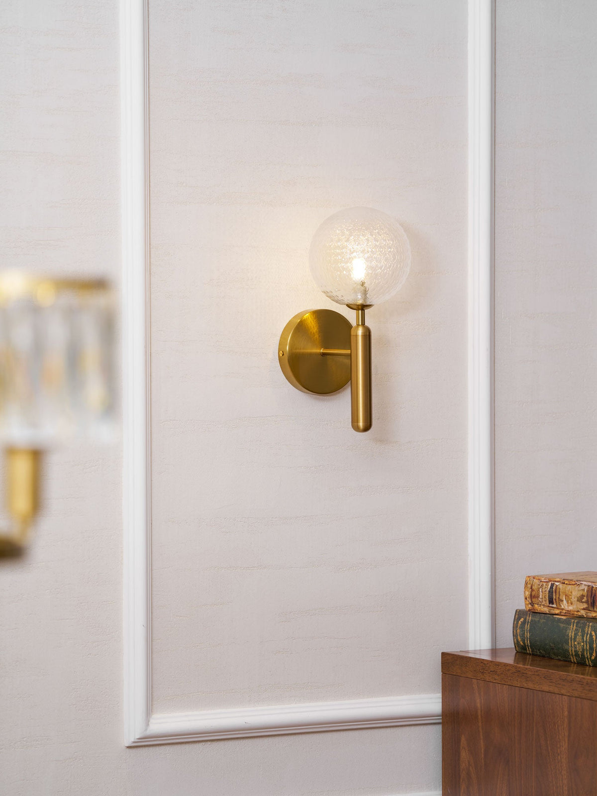 Near a stack of books and a wood cabinet, this brass Clérachic wall light's dimpled clear glass globe glows on a panelled wall.