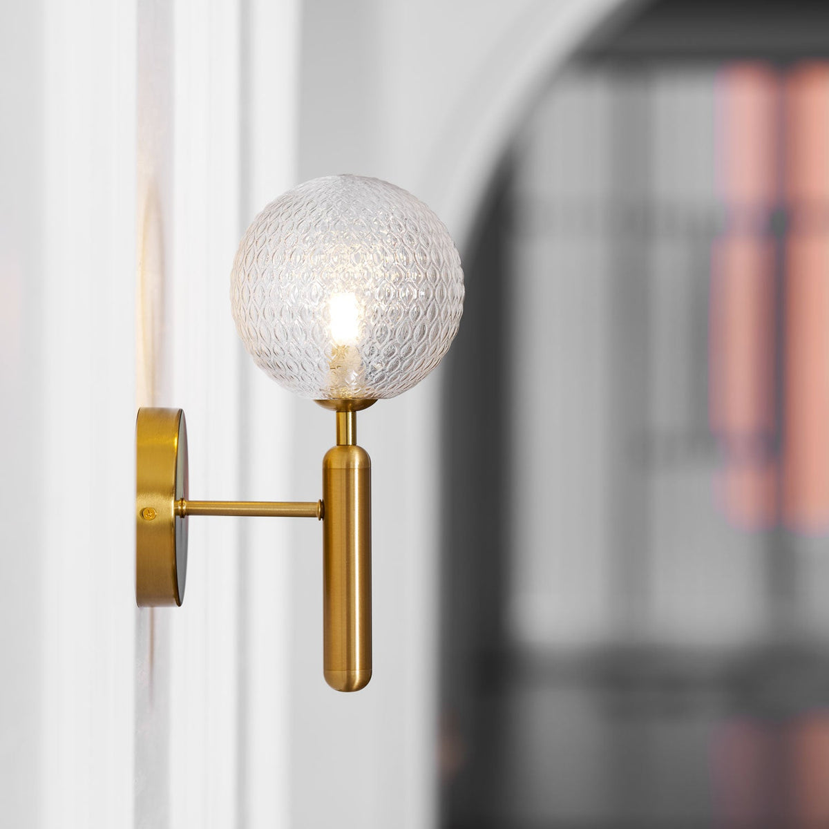 Beside a white door with a dark iron grille beyond, this brass Clérachic wall light's dimpled clear glass globe glows softly.